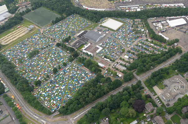 Aerial view of a densely packed camping area with rows of colorful tents next to event stages and parking lots, surrounded by greenery and roadways.