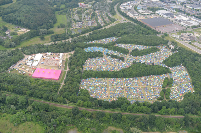 Aerial view of a large outdoor festival site with numerous tents organized in rows, surrounded by greenery, with roads, buildings, and parking lots in the vicinity.