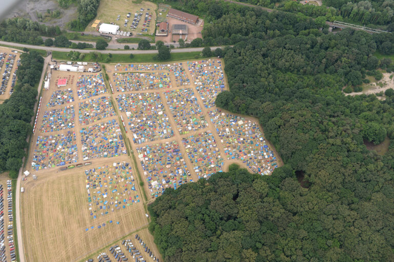 Aerial view of a large, organized camping area with numerous tents set up in rows surrounded by trees and adjacent to parking lots.