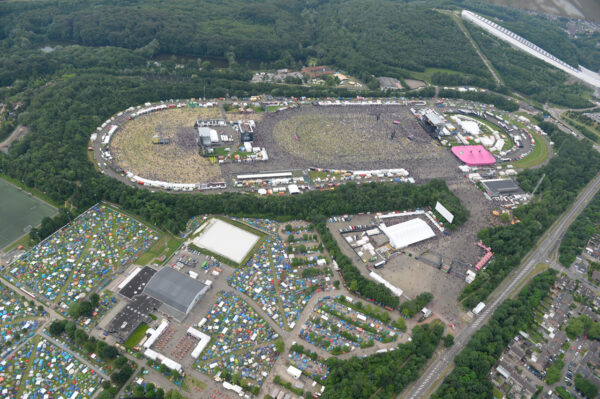 Aerial view of a large outdoor festival with numerous attendees, stages, tents, and surrounding greenery.