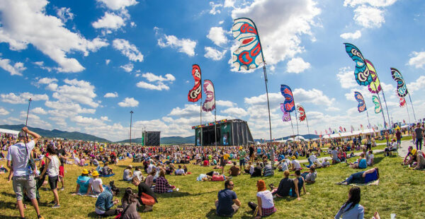 A music festival scene with a crowd of people sitting on the grass under a clear blue sky, with colorful banners fluttering in the wind and a stage in the background.