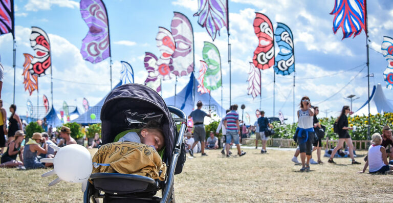 A child sleeping in a stroller at a lively outdoor festival with people and colorful banners in the background.