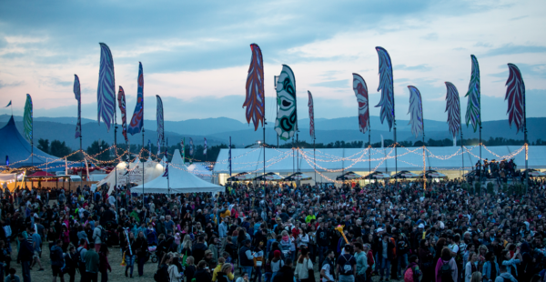 A bustling outdoor festival at dusk with a large crowd of people, colorful feather-shaped banners fluttering in the wind, and string lights above tented areas, with mountains in the background under a twilight sky.