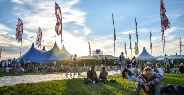 An outdoor festival scene with people sitting on the grass, walking, and socializing. Tents, colorful flags, and a setting sun in the background create a festive atmosphere.