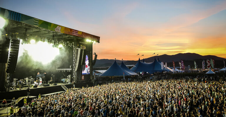 Outdoor music festival at dusk with a large crowd in front of a stage and surrounding tents, with hills in the background under a colorful sunset sky.