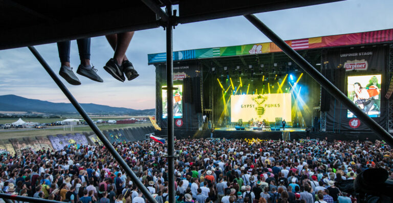A crowded outdoor music festival with a view from the stage scaffolding; the audience is focused on a band performing under a sign that says 