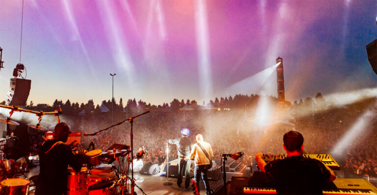 A band performing on stage at an outdoor concert, viewed from behind, with bright stage lights shining over a large crowd of spectators as the evening sky darkens.