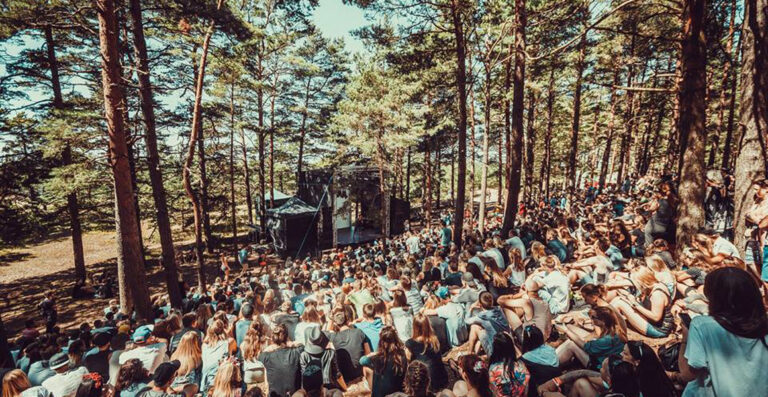 A large outdoor gathering of people sitting on a forest floor, facing a stage at a festival or event, with tall pine trees surrounding the area.