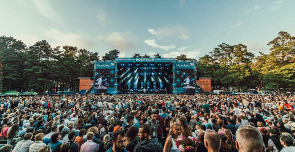 A large outdoor music festival with a dense crowd watching a performance on a stage with screens displaying the artists, surrounded by trees under a clear sky.