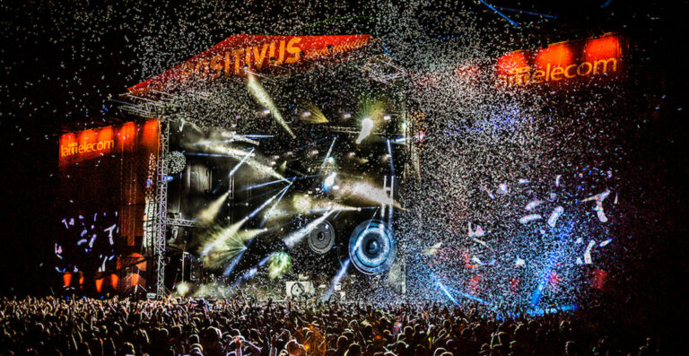 Crowd of people enjoying a concert at night with a confetti explosion, bright stage lights, and large speakers visible on a festival stage.
