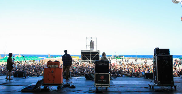 A view from behind a stage where a band is performing in front of a large outdoor audience with the sea in the background under a clear blue sky.