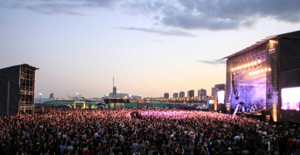 Outdoor music festival at dusk with a large crowd of people facing a stage with bright lights and screens, with city buildings on the horizon.