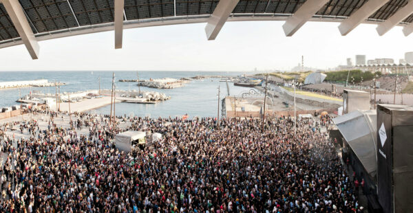 A panoramic view of a large outdoor crowd gathered for an event near the coast, with a stage on the right side and the sea in the background.