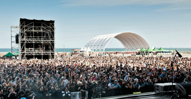 A large crowd at a beachfront music festival with a stage on the left and a curved canopy structure on the right, with the ocean in the background.
