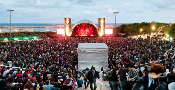 A large outdoor concert crowd gathered in front of a lit stage during dusk, with an ocean background and people leaving on the right.