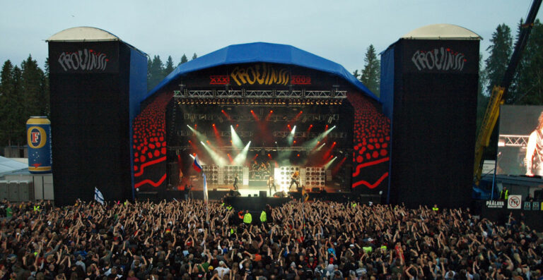 A lively outdoor music festival with a large crowd of spectators raising their hands in front of a stage with bright lights and a band performing, flanked by tall speaker towers displaying the event name 
