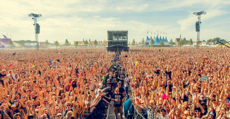 A large, enthusiastic crowd at an outdoor music festival, many people raising their hands in the air.