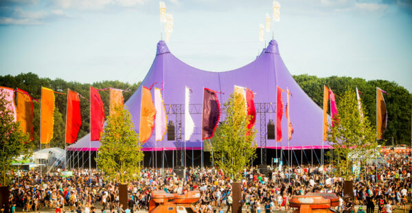 A vibrant outdoor festival scene with a large crowd of people in front of a purple circus-style big top tent and colorful vertical banners fluttering in the wind.