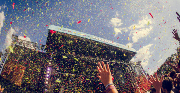 A vibrant festival scene with a crowd of people raising their hands under a sky filled with colorful confetti, with a stage structure in the background adorned with the text 