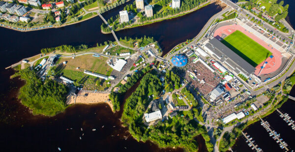 Aerial view of a bustling outdoor event next to a river, with crowds gathered around stages and tents, flanked by a stadium, green areas, residential buildings, and boats lined up along the water.