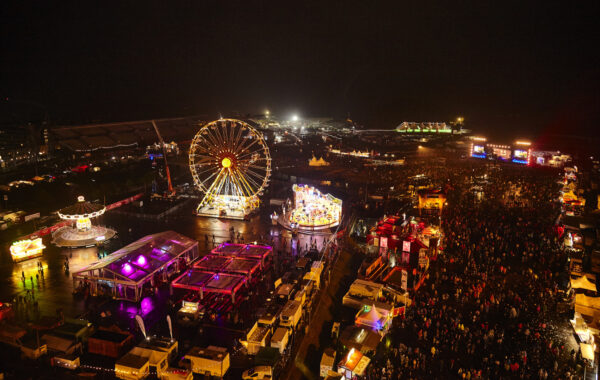 Aerial view of a bustling night-time fair with illuminated attractions including a Ferris wheel, merry-go-round, and various vendor stalls, with large crowds of people gathered.