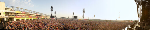 Panoramic view of a densely crowded outdoor music festival with a large audience in daylight, stage on the right, and multiple towering speaker arrays.