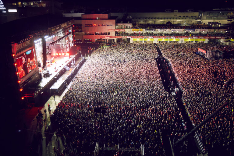 An aerial view of a large outdoor concert with a massive crowd facing a brightly-lit stage at night with screens displaying performers, surrounded by stadium seating.