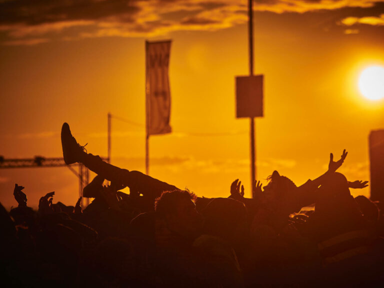 Silhouetted crowd at an outdoor event doing a crowd surf under a sunset sky.