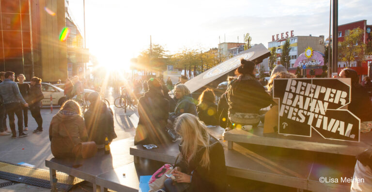 People sitting and socializing on benches and standing in a busy street environment with sunflare during sunset; signs for the Reeperbahn Festival are visible.
