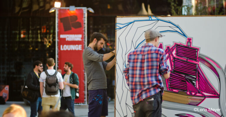 Two artists drawing on a large mural with abstract designs at an outdoor art event. People are conversing in the background next to an event lounge sign.