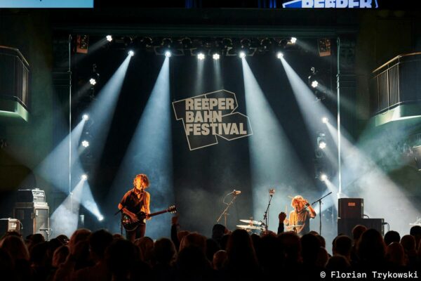 Two musicians performing on stage at the Reeperbahn Festival, with the audience in the foreground and stage lights beaming down.