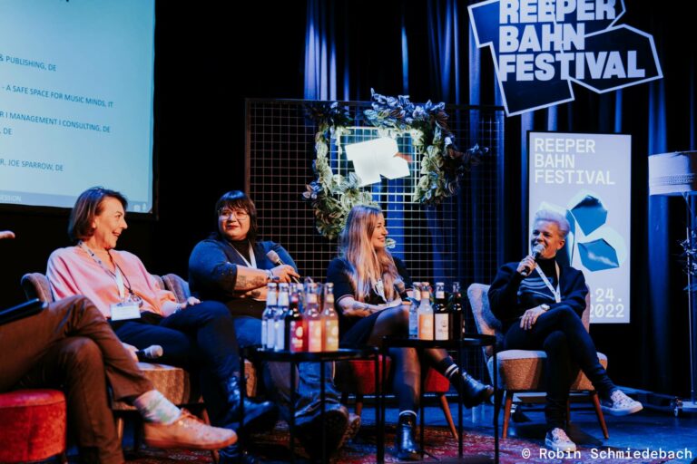 Four individuals seated in armchairs on stage at the Reeperbahn Festival during a panel discussion. The stage backdrop features the festival’s logos and a screen displaying text, with an illuminated wreath on a grid panel and water bottles on a small table in front of the participants.
