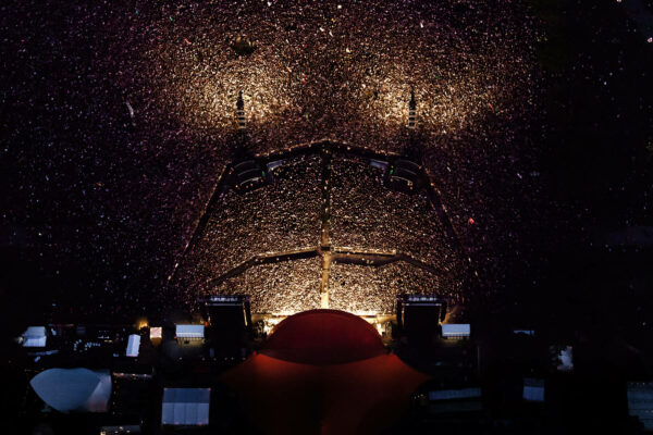 A view from the stage of a massive concert audience illuminated by numerous tiny lights, creating a starry effect against the dark night.