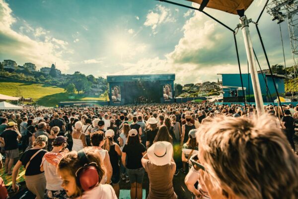 A vibrant outdoor music festival with a large crowd of attendees facing a stage with live performance screens, set against a backdrop of a sunny sky and residential hillside.