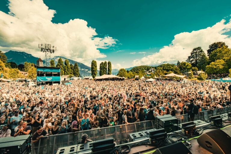 An outdoor music festival with a large, enthusiastic crowd gathered in daylight, surrounded by trees with mountains in the background, viewed from the stage area.
