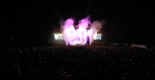 A crowded outdoor concert at night with a large audience, stage lights, and purple pyrotechnics above the stage.