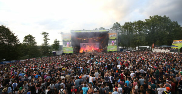 A large outdoor music concert with a dense crowd of attendees facing a stage with bright lights and large screens on either side.