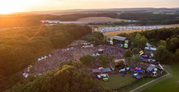 Aerial view of an outdoor music festival at sunset with a large crowd, stage, surrounding trees, and distant parking area.