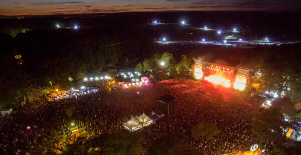 Aerial view of an outdoor music festival at night showing a large crowd gathered in front of a brightly lit stage with surrounding trees and distant lights.