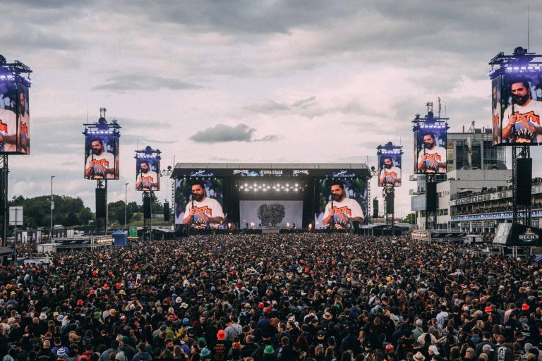 Large outdoor concert crowd facing a brightly lit stage with a man speaking on multiple large screens, under a cloudy sky.