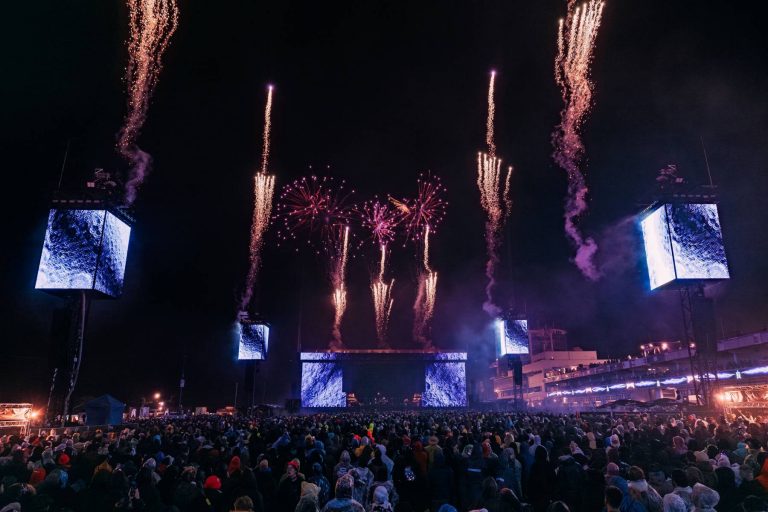 Large crowd at an outdoor night concert watching colorful fireworks burst in the sky above a stage with illuminated screens showing a moon surface image.