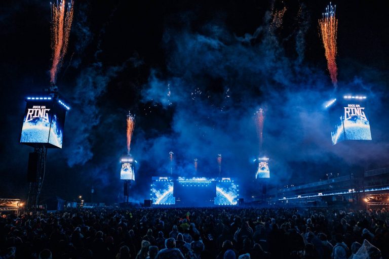 Large crowd gathered at night facing a brightly lit stage with multiple screens displaying