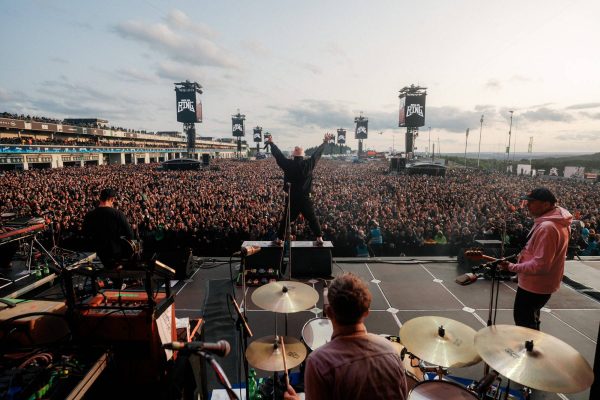 View from the stage of a band performing at a large outdoor concert with a crowd of thousands, featuring a drummer in the foreground, two guitarists on either side, and a lead singer with arms raised facing the audience under evening sky with Rock am Ring banners.
