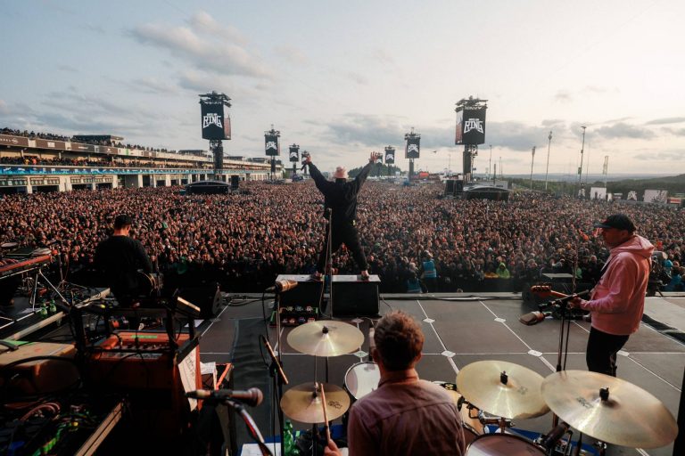 View from the stage of a band performing at a large outdoor concert with a crowd of thousands, featuring a drummer in the foreground, two guitarists on either side, and a lead singer with arms raised facing the audience under evening sky with Rock am Ring banners.