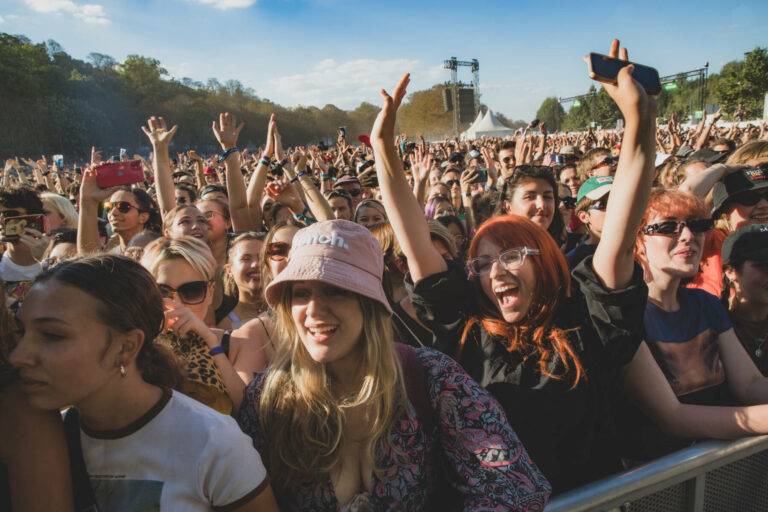 A crowd of excited concert-goers cheering and taking photos with their smartphones at an outdoor music festival.