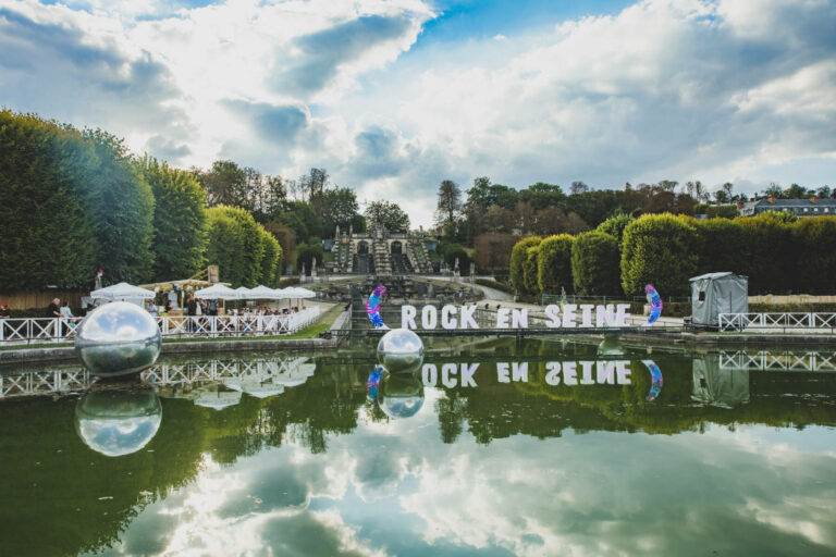 A scenic outdoor setting with "ROCK EN SEINE" signs reflected in a pond, decorative spheres on water, and a festival setup with tents and a staircase in the background under a cloudy sky.