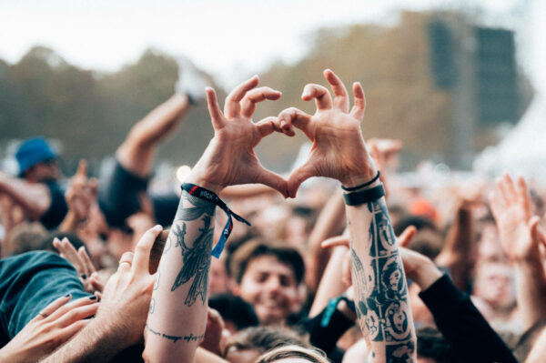 A crowd at an outdoor event with hands raised, two hands in the foreground forming a heart shape. Many attendees are wearing wristbands and some have tattooed arms.