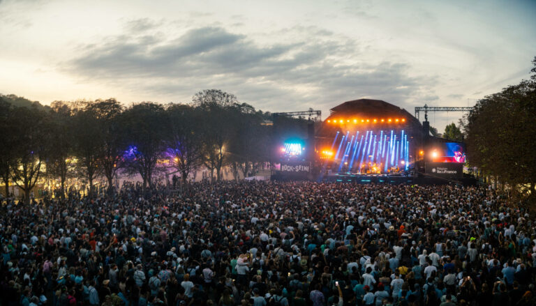 A large crowd gathered at an outdoor music festival during dusk, with a brightly lit stage in the background and the sky transitioning from daylight to evening.