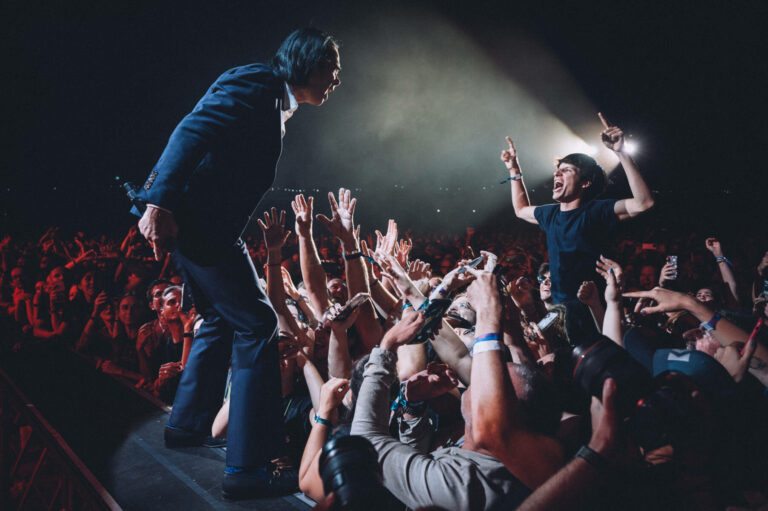Nick Cave in a dark suit leans toward an enthusiastic crowd of fans reaching out to him during a concert, with stage lights casting a dramatic beam above.