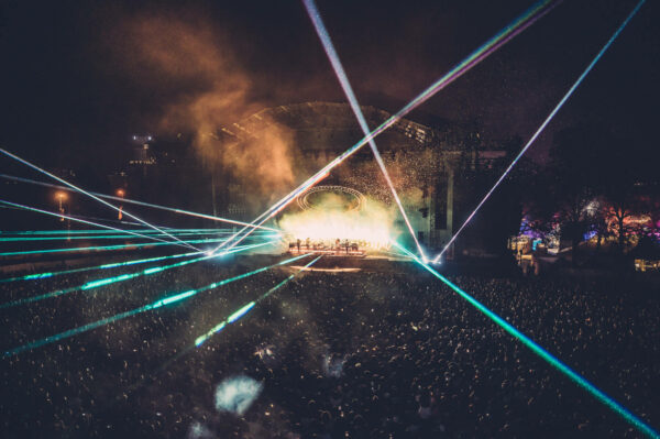 A nighttime outdoor concert scene with a large audience, stage lights, and laser beams cutting through the smoke-filled air above the crowd.
